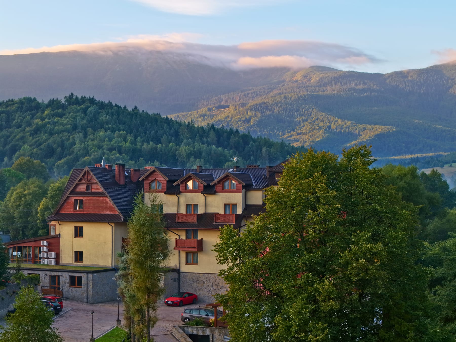 Hotel Jawor latem od strony parkingu, nad zalesionymi grzbietami chmura na Babiej Górze
