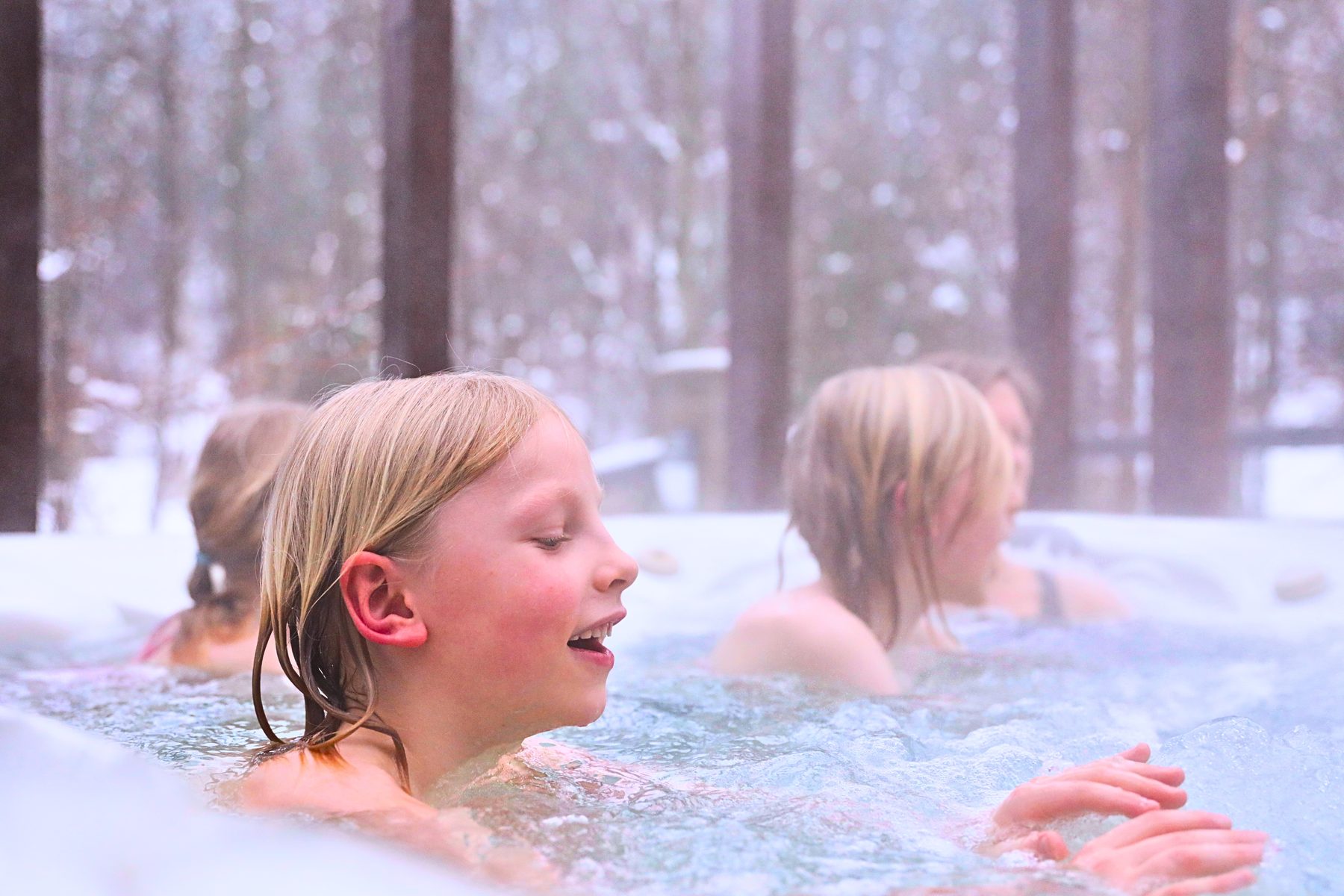 Troje dzieci w jacuzzi zewnętrznym zimą — para nad wodą i ośnieżony las za barierką