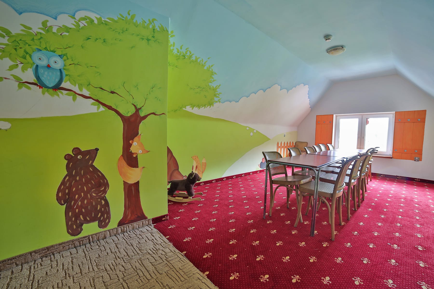 Stoły i krzesła do zajęć w kidsroomie, na ścianie malowidło z misiem, lisem i sową na drzewie