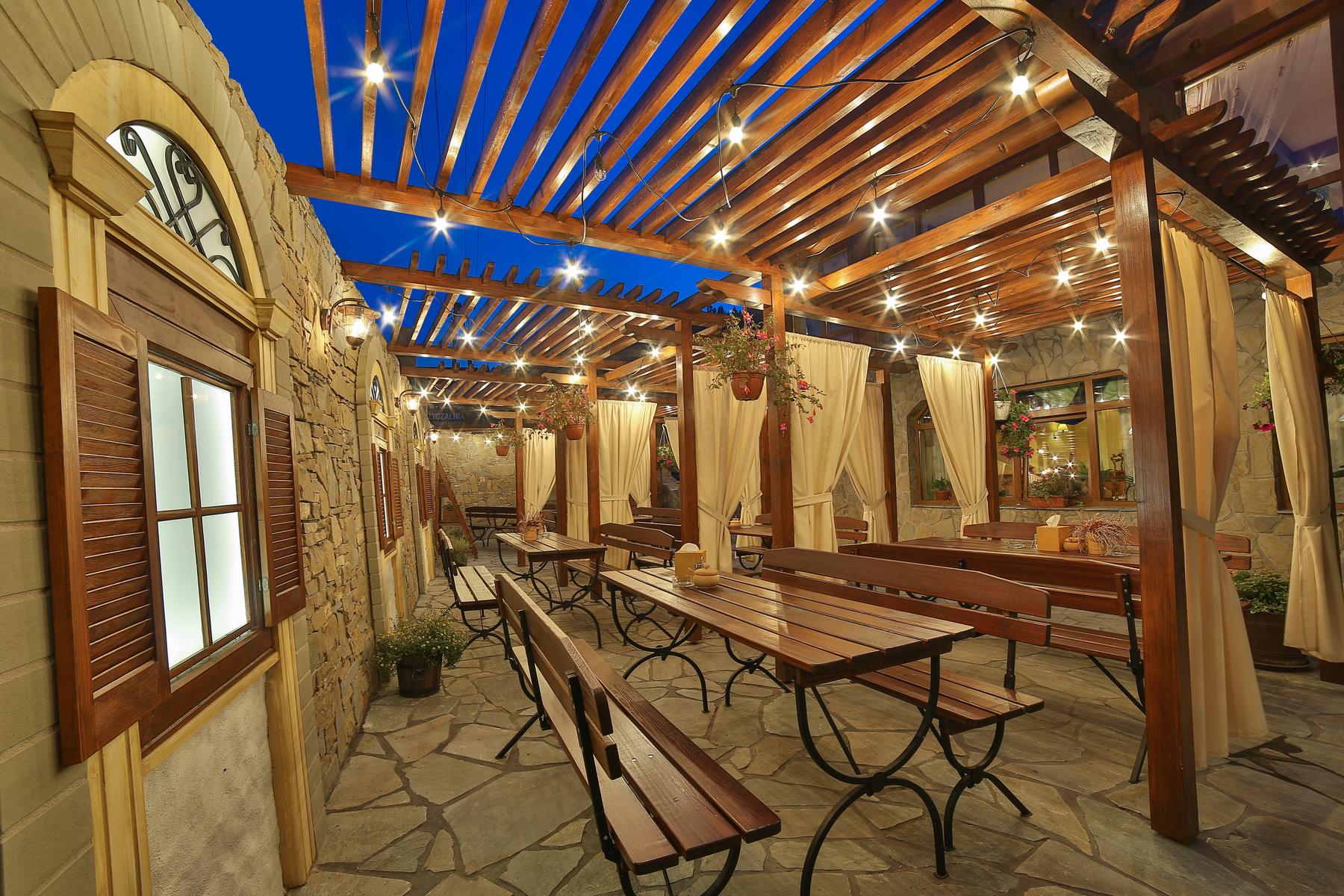 Ogródek restauracji po zmroku — pergola z lampkami i ławy biesiadne