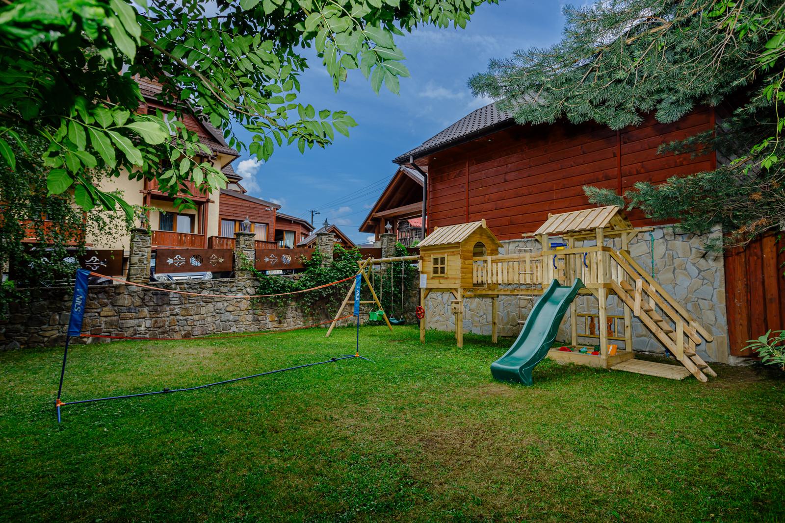 Plac zabaw przy basenie letnim — drewniany domek ze zjeżdżalnią i rozstawiona siatka do badmintona
