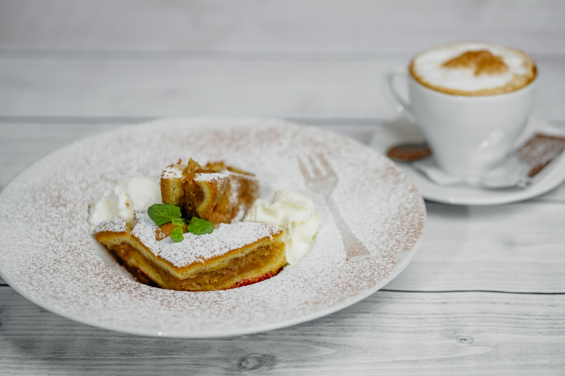 Kawałek ciasta z jabłkami i bitą śmietaną, obok filiżanka cappuccino