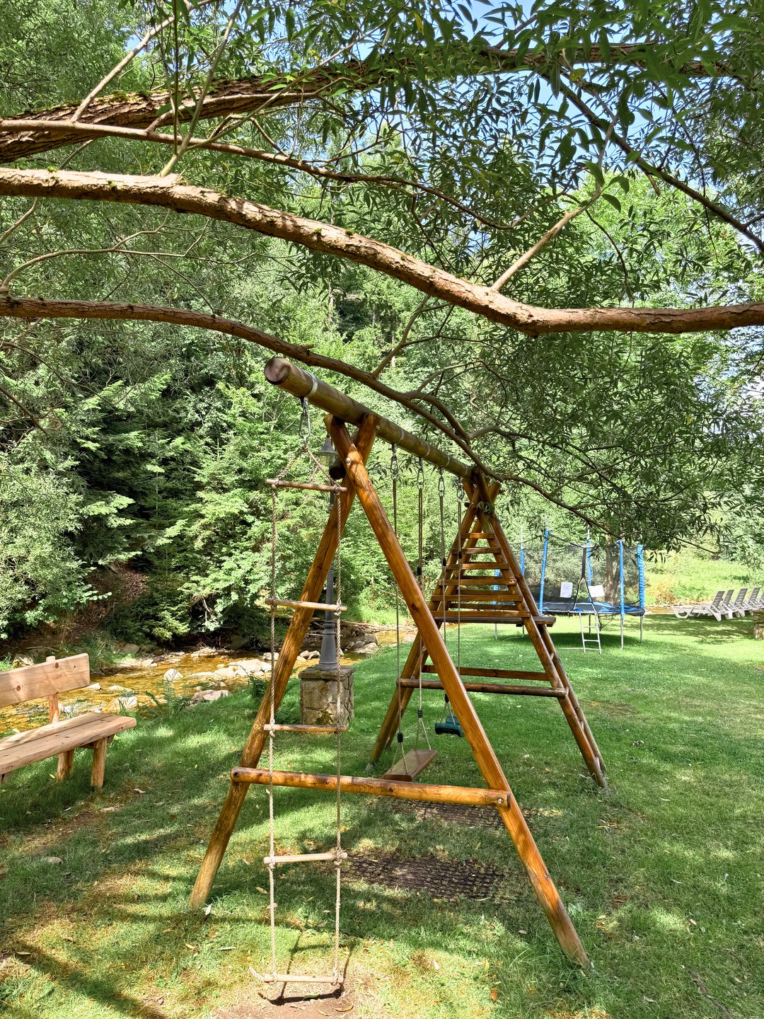 Drewniana huśtawka z drabinką linową i trampolina na terenie zielonym nad potokiem — plac zabaw przy Hotelu Jawor