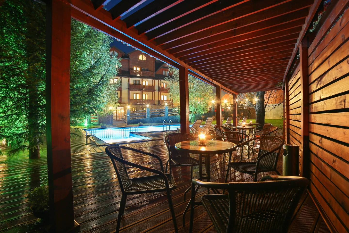 Taras pod pergolą wieczorem, w tle oświetlony basen zewnętrzny i budynek hotelu