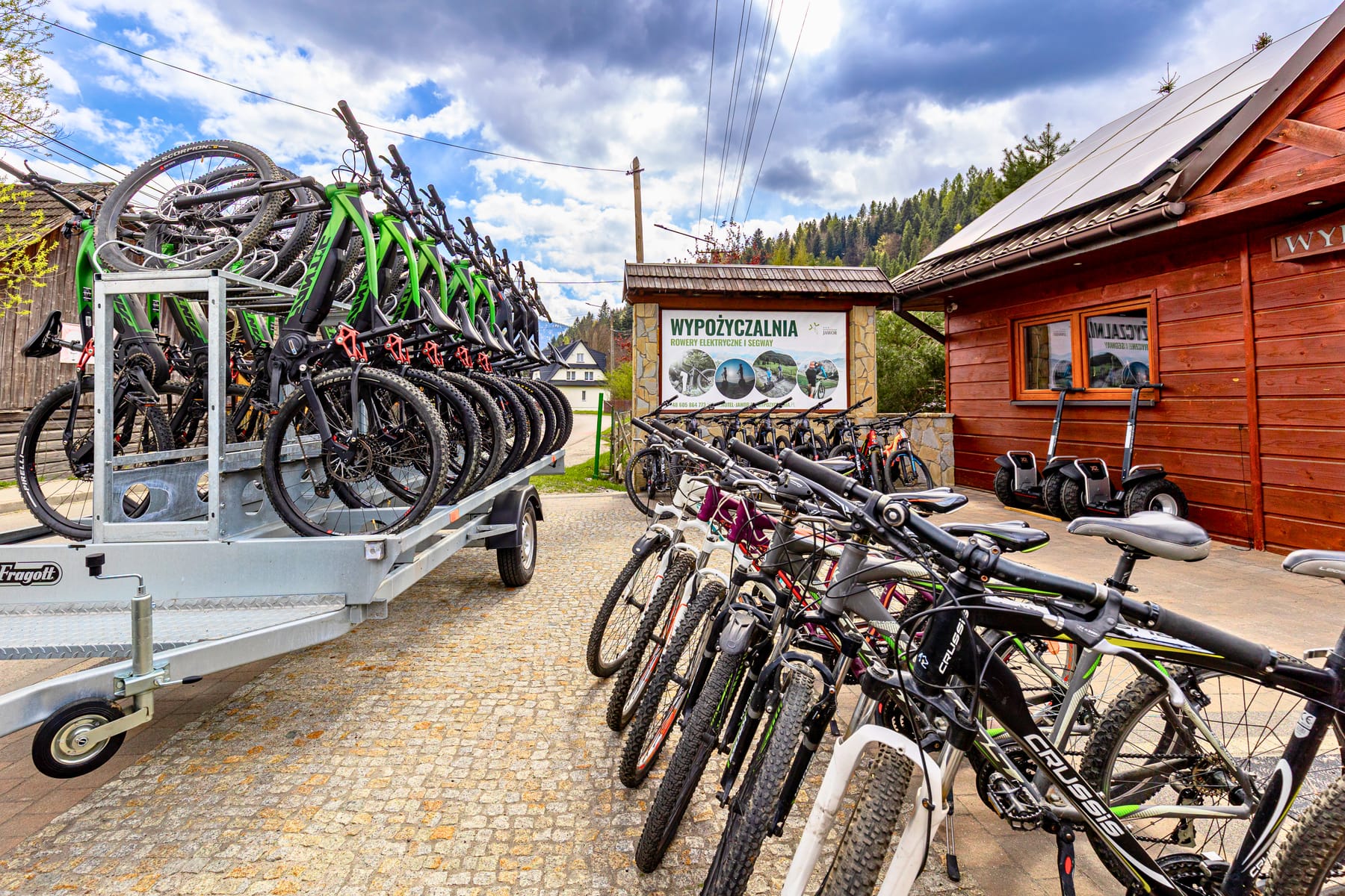 Wypożyczalnia Jawor przy hotelu — przyczepa z rowerami górskimi, stojak z e-bike'ami i segwaye przed drewnianym budynkiem