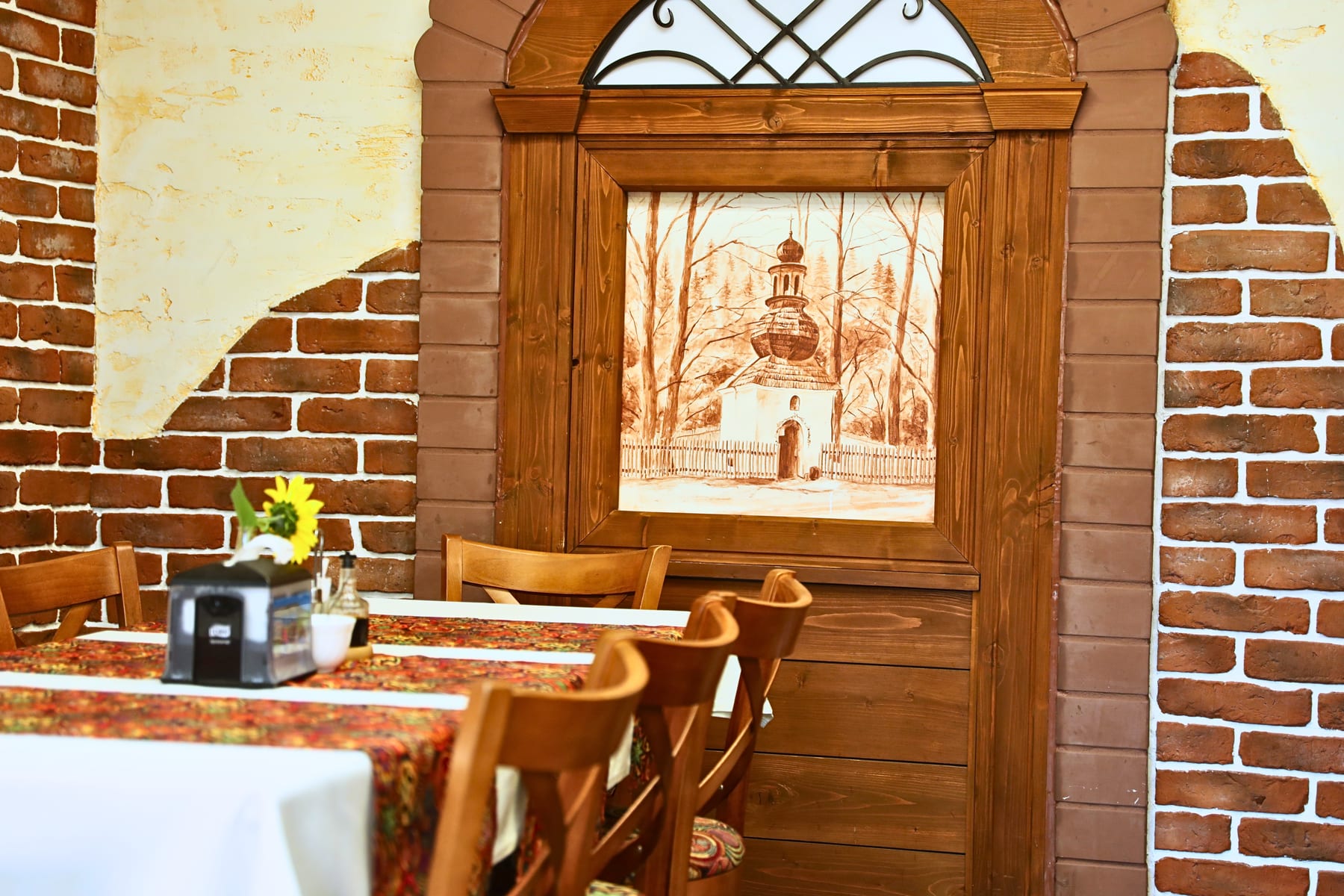 Obraz z drewnianym kościółkiem w rzeźbionej ramie na ceglanej ścianie restauracji